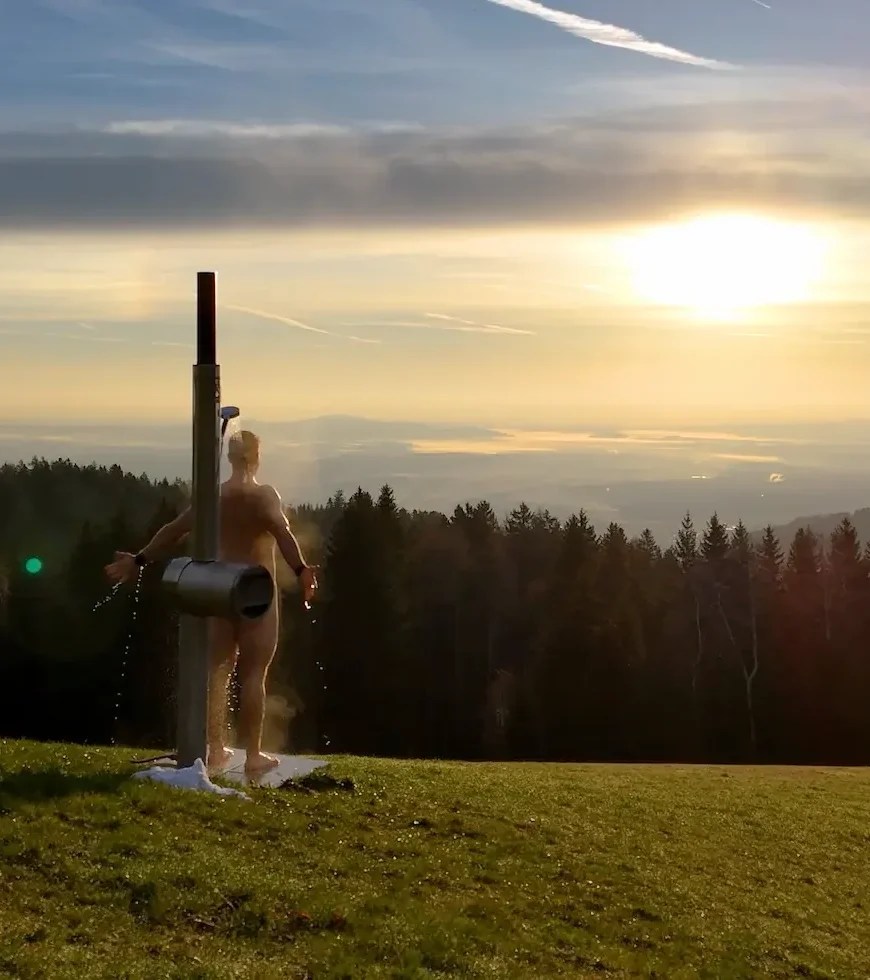 Person showering outdoors with the Feuerwasser wood-fired shower against a sunset forest view