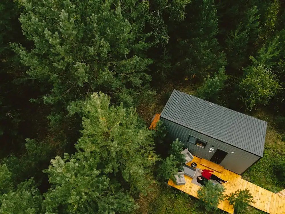 Aerial view of a small modern cabin surrounded by tall green trees, featuring a wooden deck with outdoor seating.