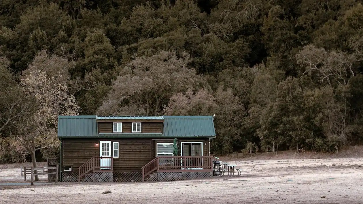 A small, rustic wooden cabin with a green metal roof, surrounded by trees and open land.